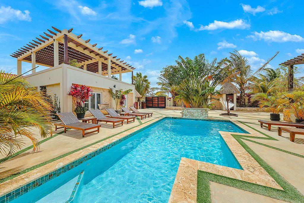 Swimming pool with wooden sun loungers around it, a pergola-topped building on the left and palm trees under a blue sky.