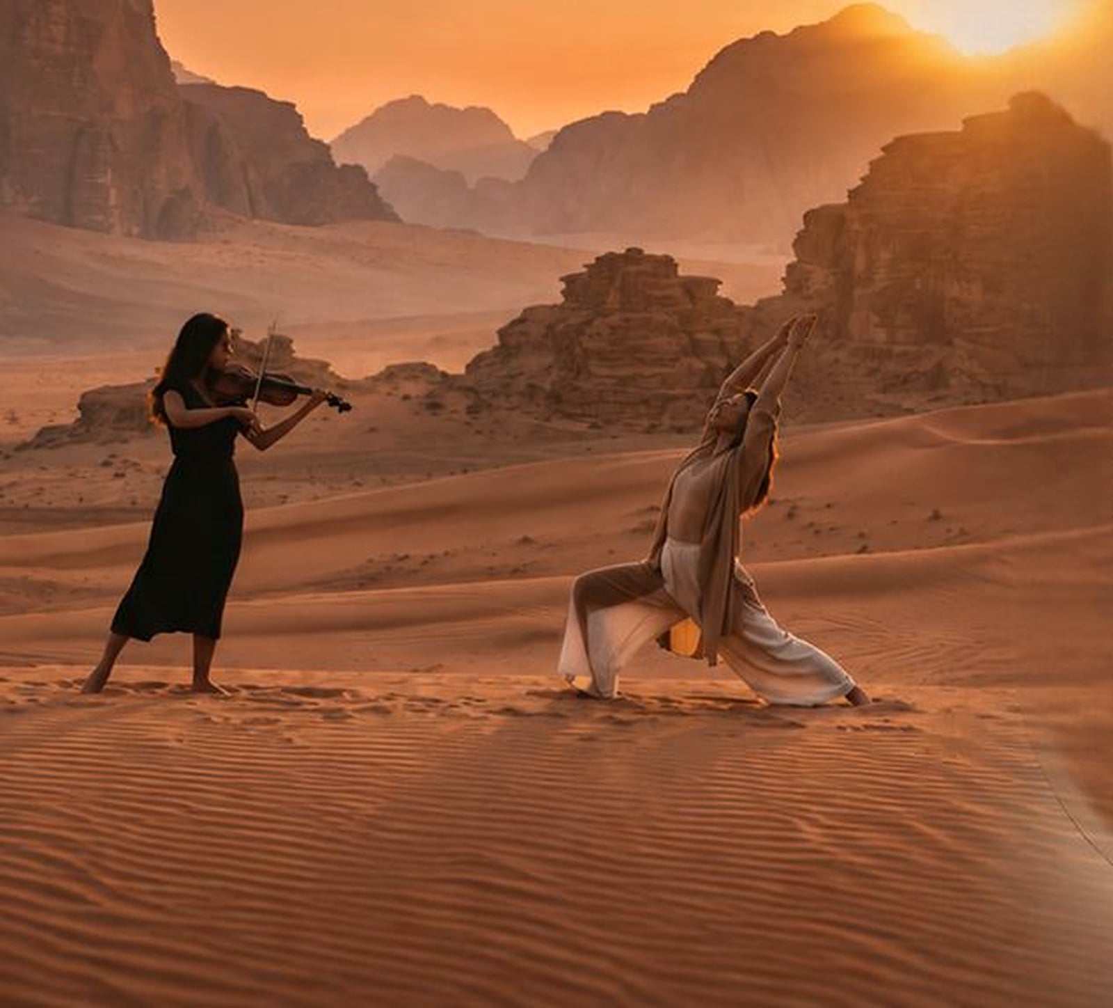 A violinist plays beside a person in a yoga pose on the dunes of Wadi Rum at sunset